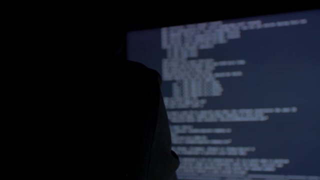 Hacker in hood cracking code using computers in dark room