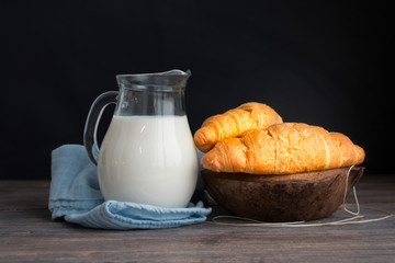 Croissants and jug of milk on wooden table against black backgro