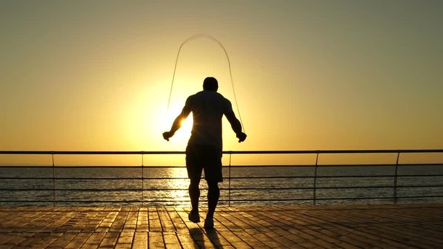 Back view man silhouette jumping with skipping rope outside at the pier on sunset