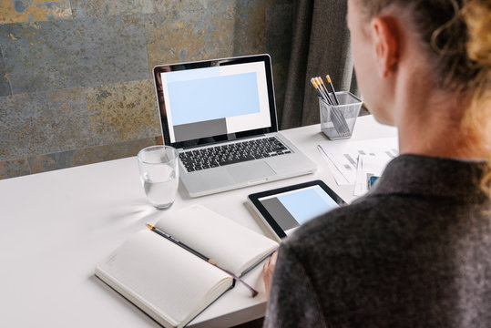 Back view. Business woman in gray shirt sits at white desk. On table is 
laptop with blue screen,notebook,digital tablet,glass of water and 
pencil holders.