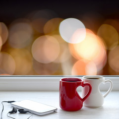 Two cups of coffee, smartphone and headphones on a windowsill. In the background, the bright lights of the city