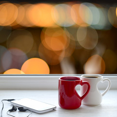 Two cups of coffee, smartphone and headphones on a windowsill. In the background, the bright lights of the city