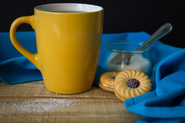 Yellow mug with hot coffee, two cookies, sugar and blue kitchen towel on wooden table. Morning and breakfast concept
