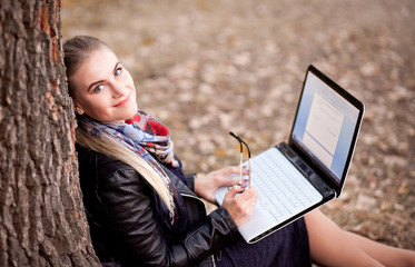 A girl student with a laptop outdoors in the park sitting under a tree in autumn