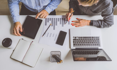 Top view of businessman and businesswoman sitting at white desk and do work together.Woman makes note on chart,man uses tablet computer.On table are laptop,notebook,smartphone,pencil holders and paper