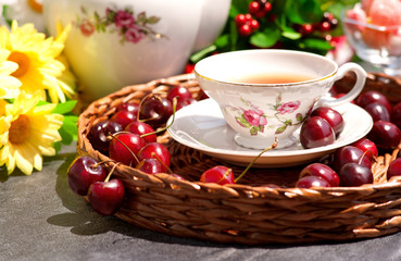 Изящная чашка чая в окружении ягод вишни. Вишневый чай стоит на плетеном круглом подносе на фоне цветов. Солнечные лучи освещают чайную пару.