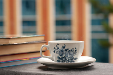 Горячий чай в чашке с синим рисунком. От чая идет струя пара. Чашка чая стоит на подоконнике балкона. Стопка книг создает домашнюю обстановку.