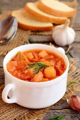 Cabbage soup with meat and vegetables in a bowl, spoon, garlic, pieces of bread on old wooden background. Soup cooked with meat, potatoes, cabbage, carrots garlic, onion and tomato. Closeup