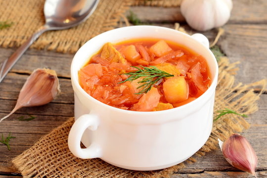 Soup with meat and vegetables in a bowl, spoon, garlic on old wooden background. Soup cooked with meat, potatoes, cabbage, carrots garlic, onion and tomato. Closeup
