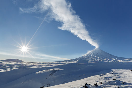 Winter view of eruption active Klyuchevskoy Volcano (Klyuchevskaya Sopka) and sun rays on beautiful blue sky on frosty day. Beautiful volcanic landscape of Kamchatka Peninsula in Russian Far East.