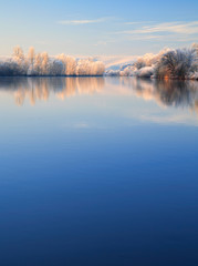 Fototapeta premium Winter Landscape Reflecting in River at Sunrise, Trees covered by hoarfrost and snow, blue sky, space for text