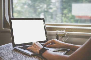 Close-up laptop with blank screen on table in train compartment. Women's hands on keyboard. On the table stands a cup of tea in the cup holder. In the background window. Girl using gadget. Mock up.