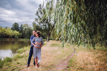 happy pregnant woman with husband standing together near the lake ina autumn sunny day. Pregnacy in long dress. Future daddy in jeans and blue shirt.