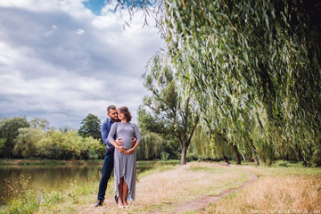Naklejka na ściany i meble happy pregnant woman with husband standing together near the lake ina autumn sunny day. Pregnacy in long dress. Future daddy in jeans and blue shirt.