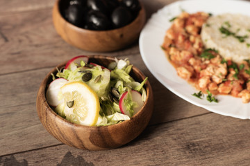Green iceberg lettuce salad with cucumbers, radishes, sunflower seeds, lemon and parmesan. Behind, in defokus chicken with rice, olives. Rustic wooden background.