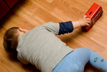 Lonely two-year baby is laying on a kitchen floor and playing with red toy   bus - 124884919