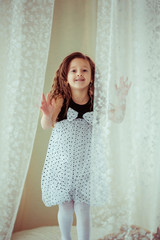 Fototapeta premium Little curly girl in spotted dress stands behind the white curta
