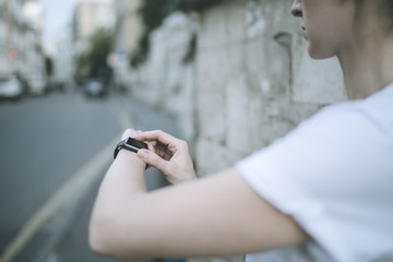 Close-up of woman's hand with smart watch. Girl in white T-shirt standing on city street and uses smart watches. A young woman uses a digital gadget.In the background is blurred road and stone wall.