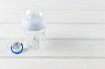 Glass baby bottle and baby pacifier on white wooden background