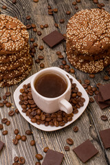 Small white cup of coffee, roasted coffee beans, cookies with sesame seeds, slices of chocolate on wooden background