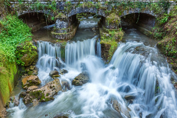 Fototapeta na wymiar Small waterfall at mountain river under the old bridge in Asturias, Spain. Waterfalls are commonly formed in the upper course of the river. 