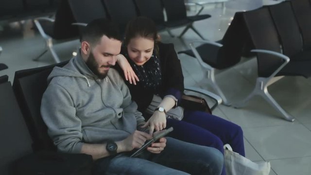 The guy and the girl in casual clothing looking at the tablet sitting on a black chairs in an airport waiting hall.