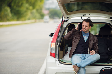 Calling friends. Happy young man talking mobile phone and sitting on a car trunk