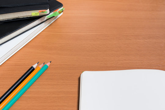 Working space with closed old notebooks with colored bookmarks and one open notebook. Three  ready to use pencils in the left corner and free space in the right corner of the image.