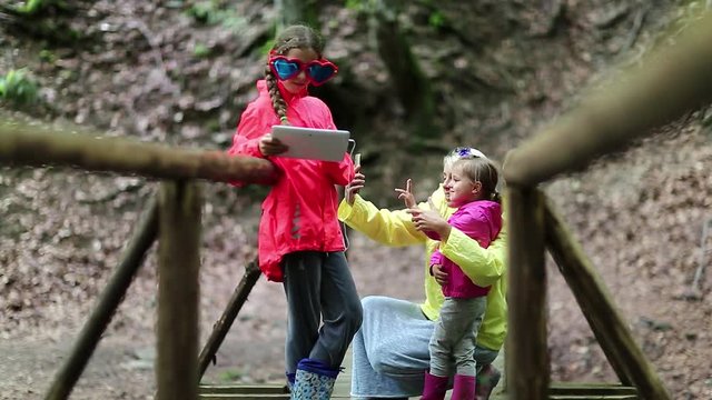 Mother and two daughters with smartphone and tablet computer on the nature. Family with electronic devices on the wooden bridge in the forest. Women in color windcheaters with phone and tablet pc