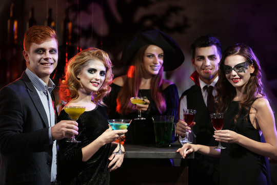 Young people in different costumes drinking cocktails at Halloween party