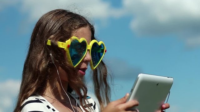 Attractive girl talks via tablet computer. Merry girl in big sunglasses in the shape of hearts communicates via tablet pc. Female in glasses with white tablet pc. Girl communicates through skype