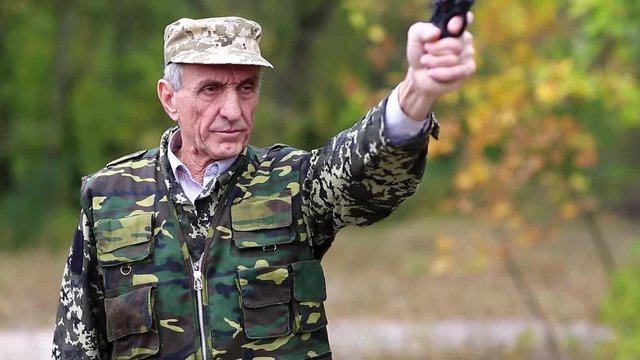 Senior man in military uniform shoots a revolver. Man with black gun. Retired officer at shooting range. Senior man in military uniform shoots a pistol in forest
