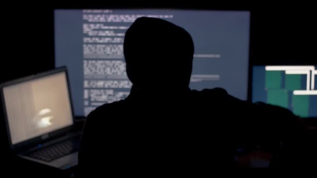Hacker in hood cracking code using computers in dark room