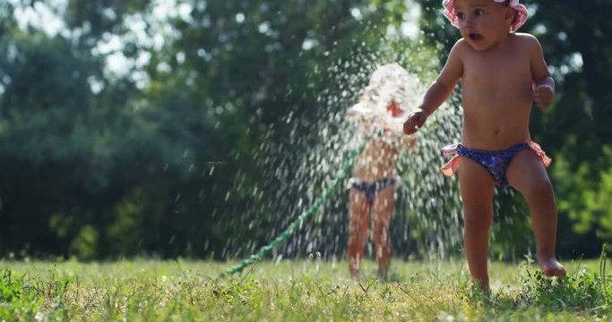 On a sunny hot day, a baby girl playing with water with her friend and sister and has fun because her friend is afraid to get wet with water. Little girl is happy because her friend flees from water