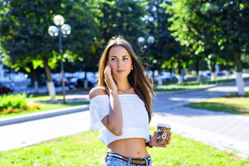 Fototapeta na wymiar Woman talking on the phone, holding a coffee or tea, beautiful brunette girl, standing in the park, summer, white blouse, long hair, sensual look, concept of student, fashion lifestyle, rest in .