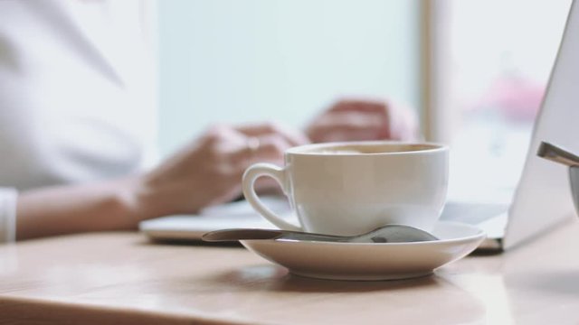 Cropped blurred female hands typing at laptop and taking drink from coffee cup near her.