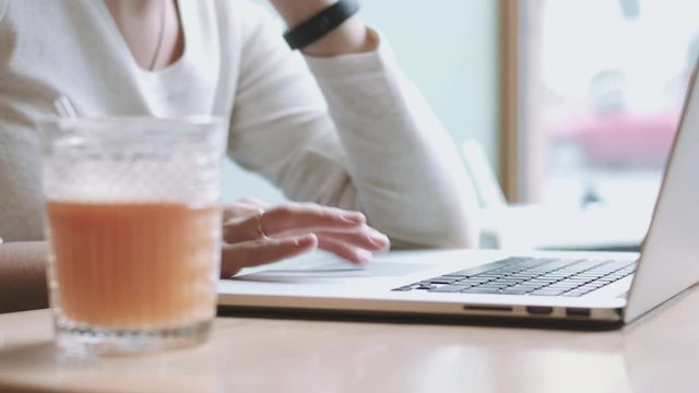 Cropped faceless woman sitting at table using laptop and drinking juice from faceted glass near her.