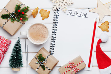 Cup of hot cocoa, holiday decorations, gift, present, miniature fir tree and notebook with to do list on white wooden table from above. Christmas or winter planning concept. Flat lay style.