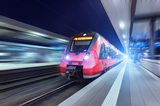 Fototapeta Modern high speed red passenger train moving through railway station at night. Railway station in Nuremberg. Railroad with motion blur effect. Industrial concept . Railroad travel, railway tourism