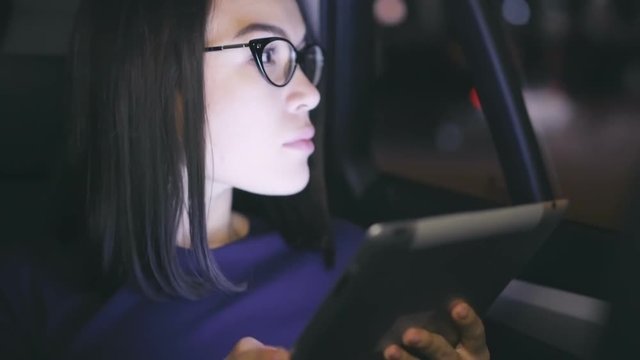Closeup of young and beautiful Asian woman in glasses looking seriously at her digital tablet screen and taking thoughtful glance on the city while being driven to the place of destination