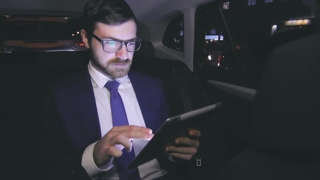 Young and handsome Caucasian businessman in suit with tie and glasses sitting in car backseat, reading news on digital tablet and taking a rapid glance on the city while being driven