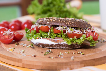 A healthy meal of whole wheat dark (brown) rye bread, curd cream cheese, radish, alfalfa sprouts, ham, cherry tomatoes and green salad on a wooden cutting board. Delicious breakfast full of vitamins.