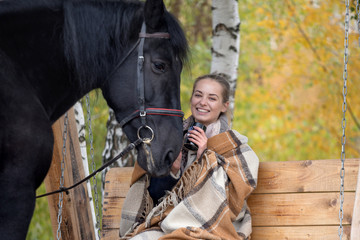 girl in a plaid with a black horse in the autumn under a birch tree on a bench