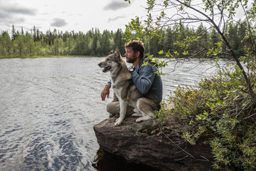 Handsome man and dog are sitting on the stone near of lake and looking into the distance