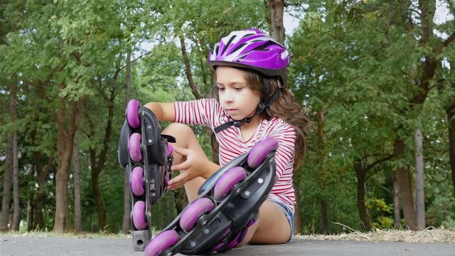 Cute little girl with protection hat putting on rollers in park alley