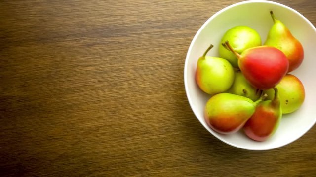Gliding overhead shot of small green and red pears in a white bowl against wood background with copyspace