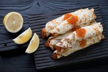 Pancakes stuffed with red caviar on a black wooden serving board