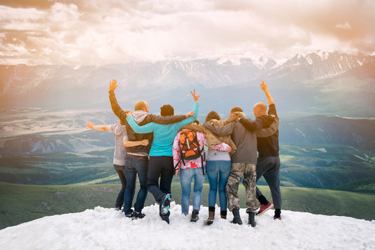 Group Of Friends Are Happy That Climbed To The Top  The Mountain. They Hug And Jump. Looking Into The Distance With Back  The Camera.