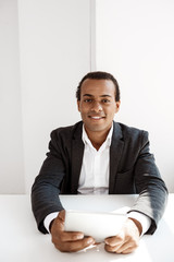 Young successful african businessman smiling, holding tablet, sitting at workplace.