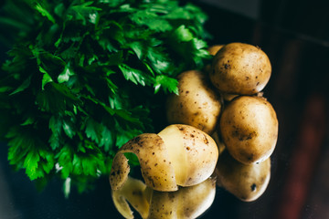 A bunch of parsley and raw unpeeled potatoes are on a black background and reflects in the mirror.